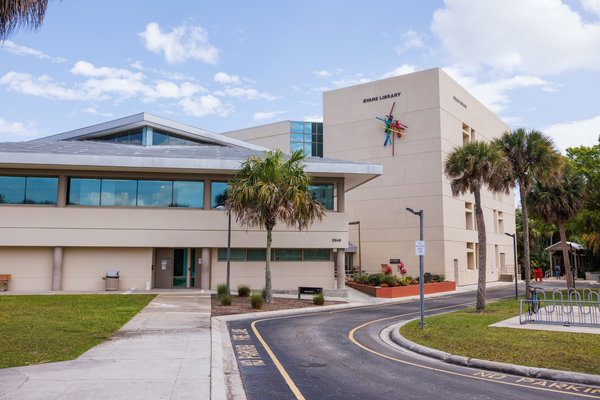 The front of Evans Library, a four-story building, with some palm trees. Blue sky with wispy white clouds. Sidewalk and roadway leading up to and pas the library.