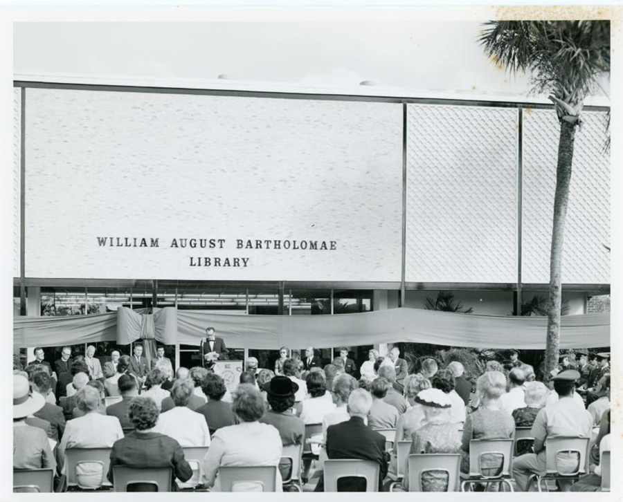 Founding President Jerry Keuper dedicates teh William August Bartholomae Library (now the Keuper Building) at a BEC podium in front of a crowd of people sitting in plastic chairs in front of the entrance. A large bow wraps the building.